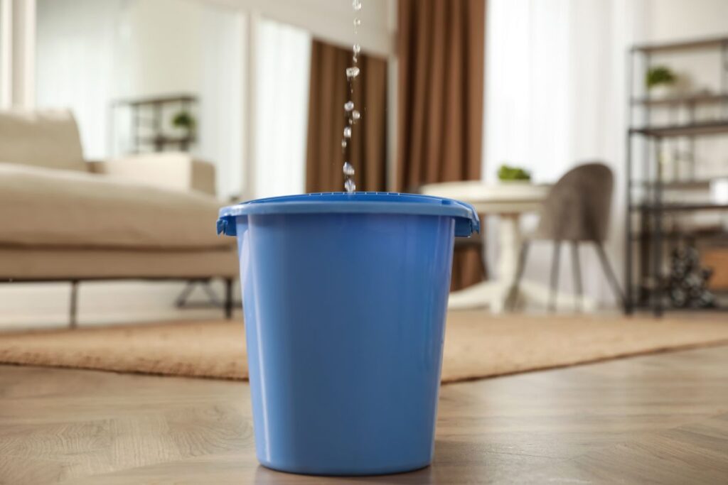 A blue bucket in the middle of a living room, catching water from a leaking roof.